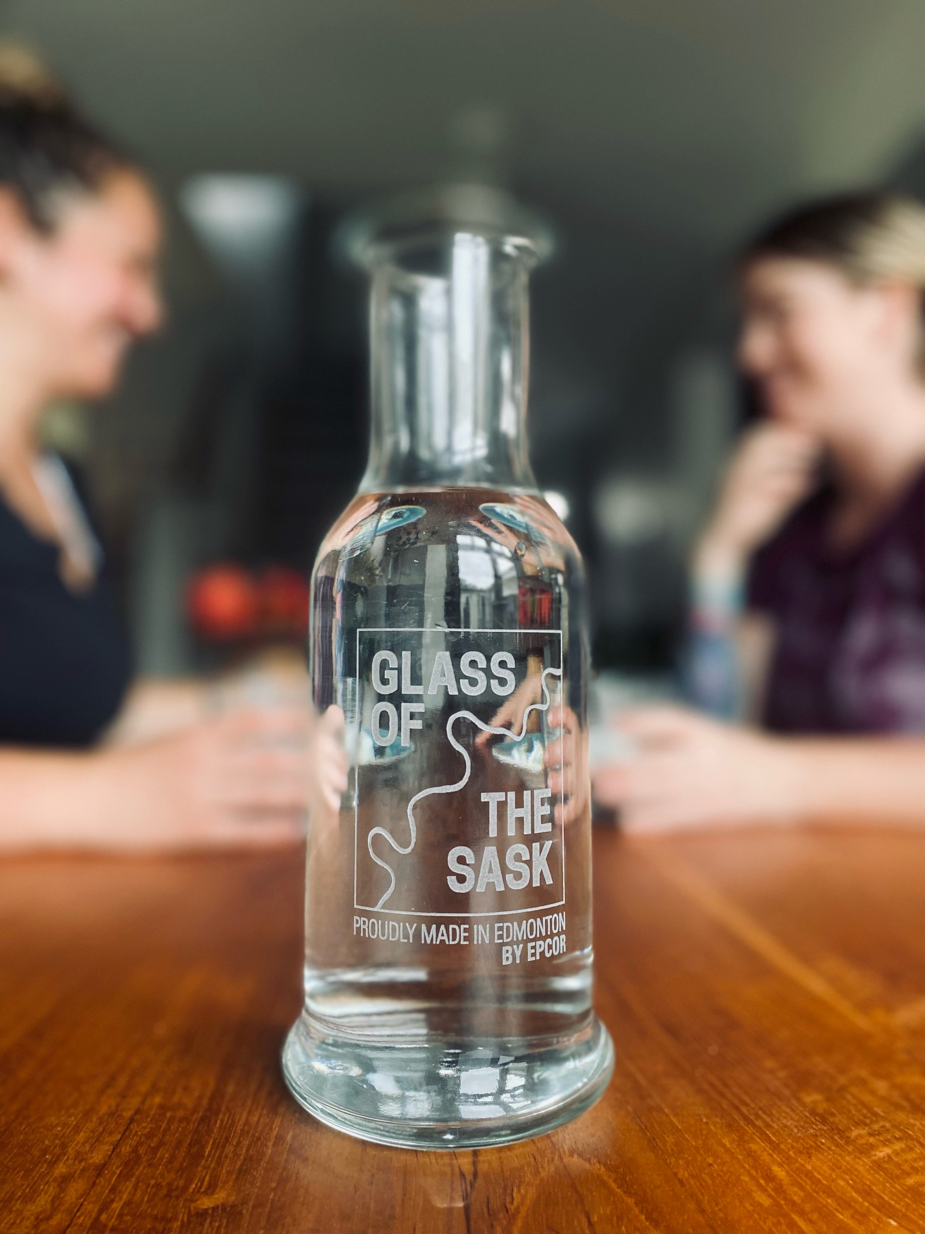 Clear glass carafe on a wooden table. Glass reads "Glass of the Sask." Two individuals are in the background, slightly blurred out, looking at each other. 